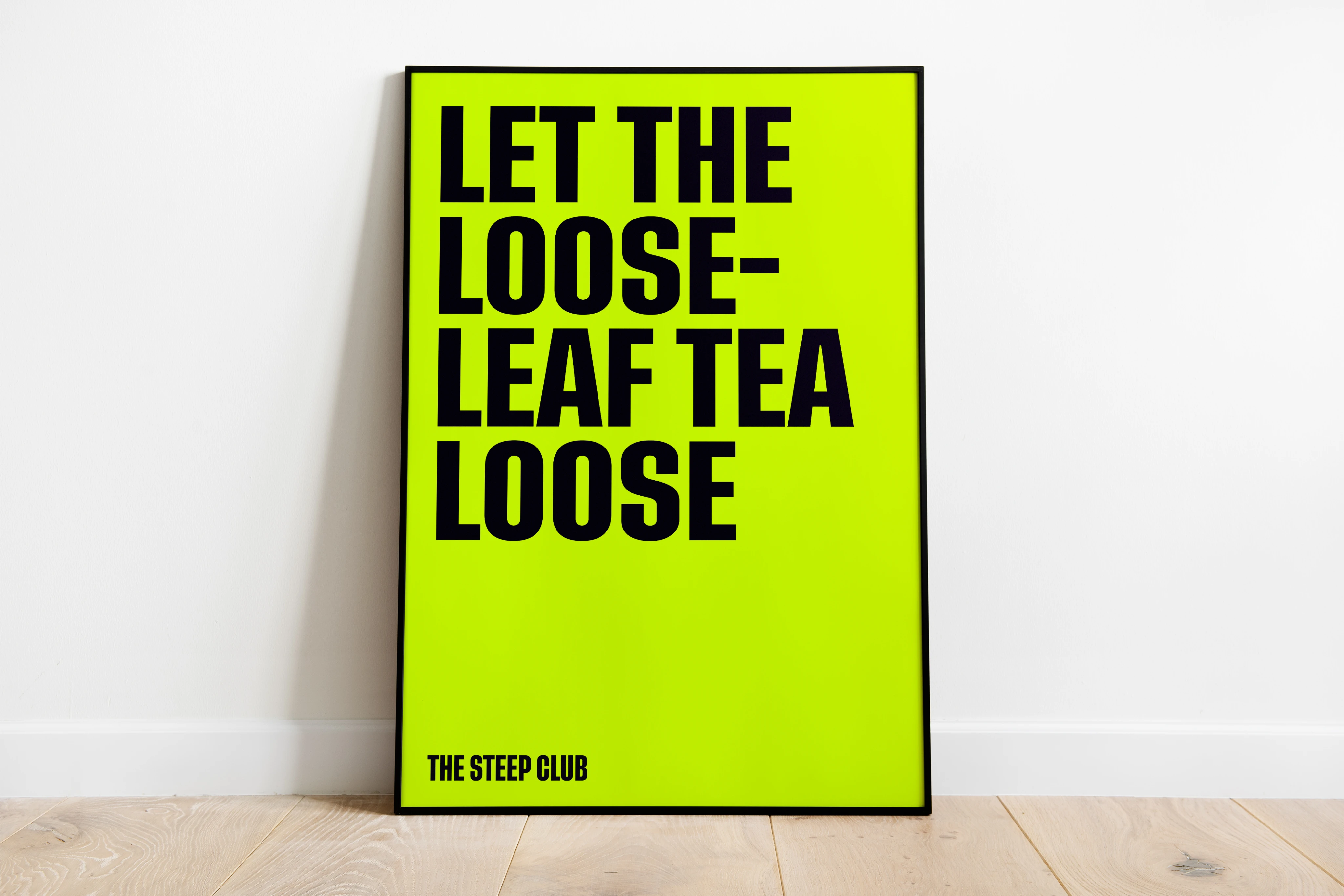 A framed acid yellow poster, “Let the loose-leaf tea loose”, leaning against a white wall on a wooden floor